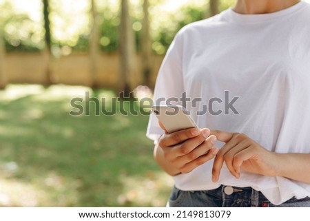 A beautiful lady is walking and doing something with a phone. We can see a park on the background and a lot of trees. She is wearing a white T-shirt. The weather is good.