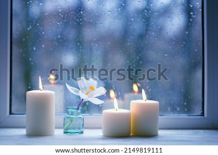 Burning candles, white flower in bottle on windowsill, abstract blurred raindrops glass background. Melancholy cozy mood. Relax, harmony, meditation, life balance concept. rainy weather