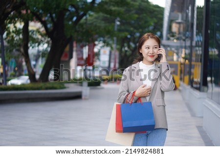 An Asian woman with a colorful shopping bag smiles happily in front of a shopping mall.