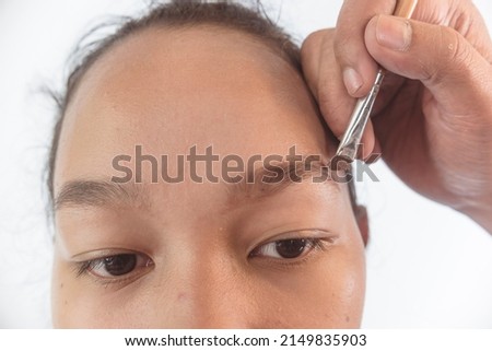Closeup of a MUA or Makeup artist using brow filler on a slanted eyeliner brush to shape the eyebrow of a young female asian model. Early stages of makeup. Royalty-Free Stock Photo #2149835903