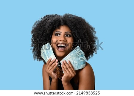 young black woman smiling holding brazilian money bills, positively surprised, space for text, person, advertising concept, isolated on blue background.