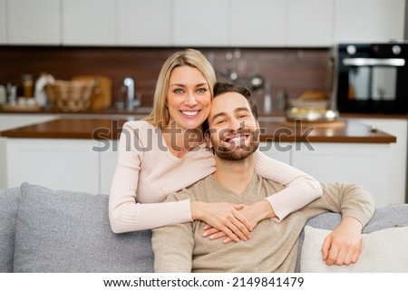 Cheerful caucasian couple in love sitting on the sofa in embrace and looking at the camera. Tenderly portrait of smiling man and his girl hugging. Love and affection concept