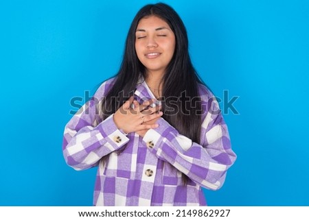 young hispanic woman wearing plaid shirt over blue background smiling with hands on chest with closed eyes and grateful gesture on face. Health concept.