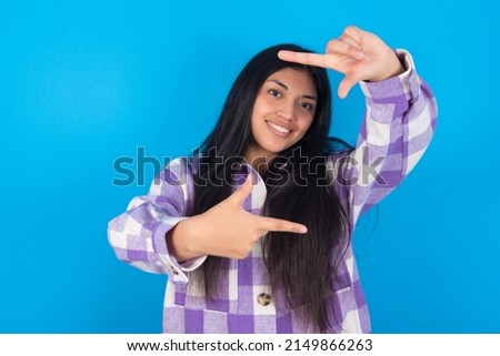 young hispanic woman wearing plaid shirt over blue background making finger frame with hands. Creativity and photography concept.