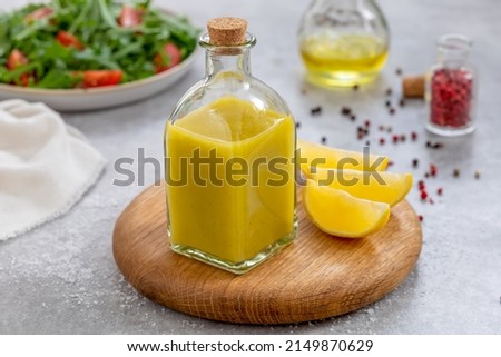Salad dressing with oil, lemon juice, salt and pepper served in glass cruet or bottle. Close up, ingredients and plate with arugula and tomato salad on background. Horizontal. Basic Vinaigrette. Royalty-Free Stock Photo #2149870629