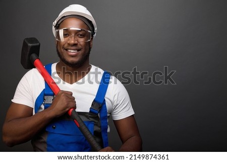 Strong black worker holds a heavy sledgehammer with a red handle. An iron hammer is carried on a strong shoulder by a courageous African-American construction worker in protective glasses and a helmet