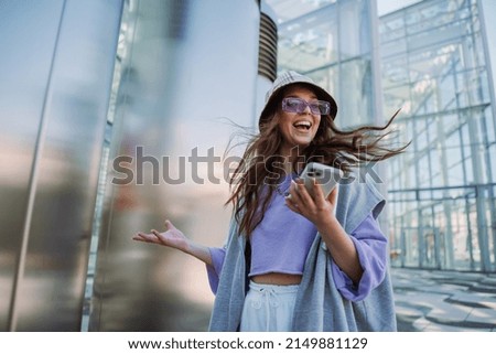 Excited young smiling woman holding and using mobile phone. Cool stylish girl at outside Royalty-Free Stock Photo #2149881129