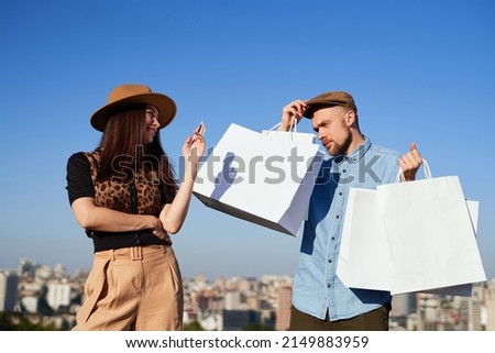 Cute shopaholic fashion styled couple: happy girl with credit card and unhappy husband in peaked cap with shopping bags in hands posing together over blue sky background. High quality photo