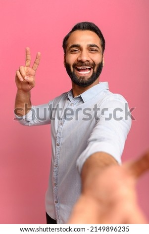 Portrait of middled-aged Indian handsome man taking selfie, looking at camera and showing V-gesture with hand, point of view of photo. Indoor studio shot isolated on pink background, vertical