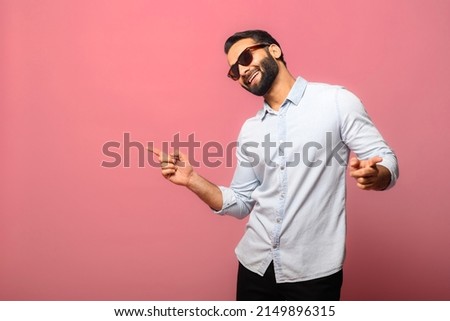 Overjoyed Indian man wearing sunglasses dancing, makes movements to music, smiles positively, being in high spirit. Carefree hispanic guy in blue jeans shirt dances isolated on pink Royalty-Free Stock Photo #2149896315