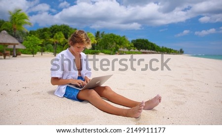 A student is engaged in self-education sitting on the beach against the background of the turquoise water. Online education concept. Young boy attending an online lesson during the COVID-19 lockdown