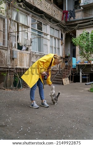 Young photographer woman in yellow raincoat playing with homeless cat on the street in old town of Tbilisi Georgia. Smiling female tourist stroking cat, touches nose. Pet lovers.
