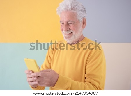 Portrait of attractive senior white haired man on colorful background while using mobile phone. Smiling bearded senior male technology addicted writes messages on cellphone