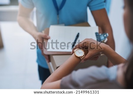 Ive been expecting this package. Cropped shot of an unidentifiable businesswoman checking the time on her watch as the courier arrives with a delivery. Royalty-Free Stock Photo #2149961331