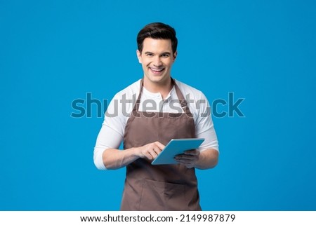 Serviced minded Caucasian male waiter with apron holding tablet in his hands while standing on blue background in light studio Royalty-Free Stock Photo #2149987879