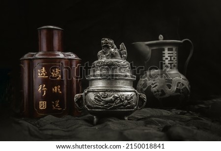 Silver antique incense burner, Chinese antique tea leaf iron storage jar (Characters chinese is Name of the tea) and Chinese antique teapot (Characters chinese is Double Happiness) on dark background. Royalty-Free Stock Photo #2150018841
