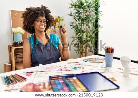 Beautiful african american woman with afro hair painting at art studio with a big smile on face, pointing with hand and finger to the side looking at the camera. 