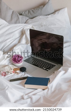Feminine still life with laptop, book, cosmetics, a jar of cream on white rumpled bed.