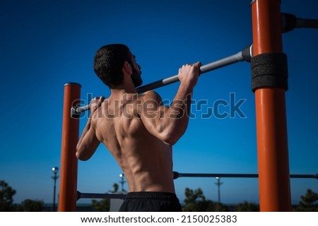 Shirtless man pulling up on horizontal bar outdoors. 
