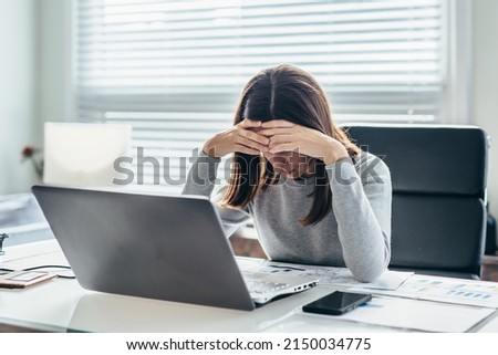 Woman sitting at table in office, leaning her head on her hand with her eyes down. Royalty-Free Stock Photo #2150034775