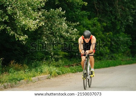 Muscular man riding his street racing bike on a road through the park. Overgrown trees by the sides. Long shot from the front.