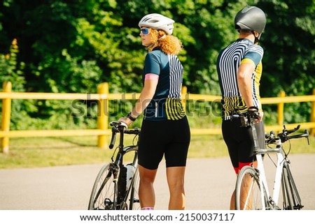 Back view of a sportive couple standing with their street bikes, blurred background. Overgrown trees behind. Side by side.