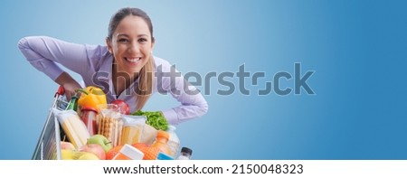 Cheerful smiling woman leaning on a full shopping cart, grocery shopping concept Royalty-Free Stock Photo #2150048323
