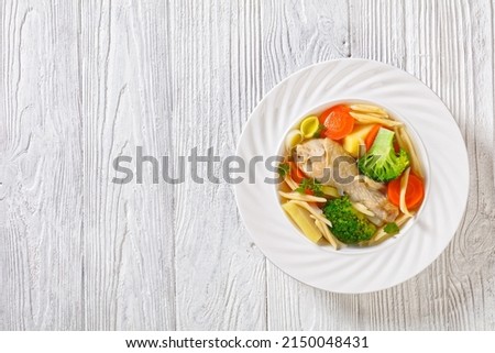 chicken spring soup with broccoli, carrots, parsnip, leek and pasta in white bowl on white textured wooden table, horizontal view from above, flat lay, free space