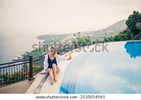 Beautiful attractive young woman sits near a large pool overlooking the sea. Vacation concept. Girl in a black swimsuit smears her skin with sunscreen