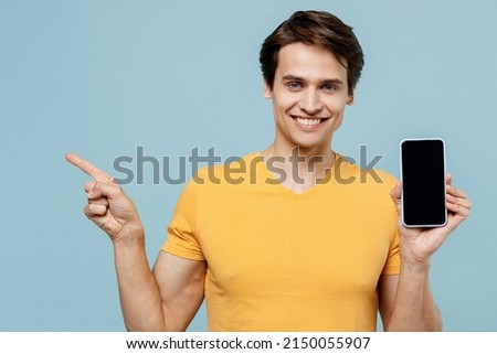 Young smiling man 20s in yellow t-shirt holding in hand use mobile cell phone with blank screen workspace area point index finge aside isolated on plain pastel light blue background studio portrait.