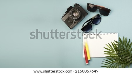 A large horizontal banner with copy space, an empty notebook with white sheets, a camera, colored markers, glasses and palm leaves on a blue background. The concept of travel, leisure, freelance