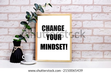 On a white table against a light brick wall, a branch of eucalyptus in a dark vase, a white cup and a light wooden frame with the text CHANGE YOUR MINDSET. Home office interior.