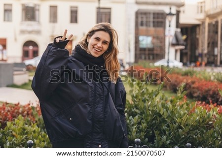Cool blonde woman in navy blue bomber shows sign v, peace and friendship with one hand , open mouth and posing outside. Happy woman walk and posing outdoor in the city. 