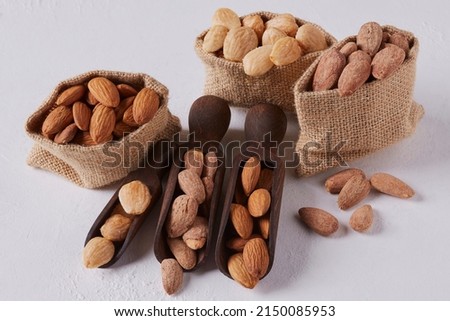 Natural, fried and roasted almonds in jute bags and black wooden spoons on white background