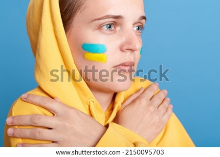 Closeup portrait of scared sad depressed woman in hoodie with Ukrainian flag on cheeks, posing isolated over blue background, keeping crossed hands on her shoulders