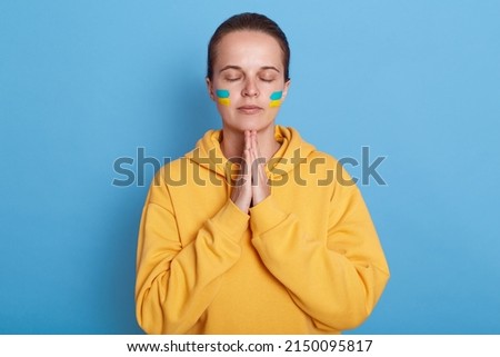 Portrait of young adult woman wearing hoodie with blue and yellow flag on cheeks isolated over blue background, praying for her motherland, keeps eyes closed and hands in praying gesture.