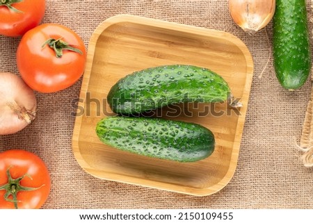 Three ripe tomatoes, two onions and two cucumbers with a bamboo plate on jute fabric, macro, top view.
