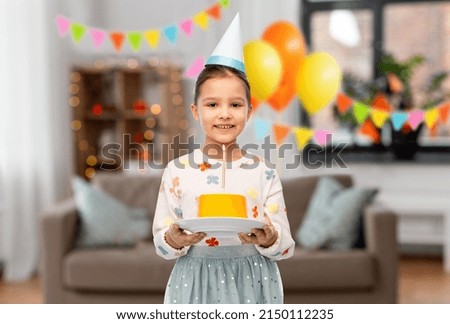 birthday, childhood and people concept - portrait of smiling little girl in party hat with cake over decorated living room background
