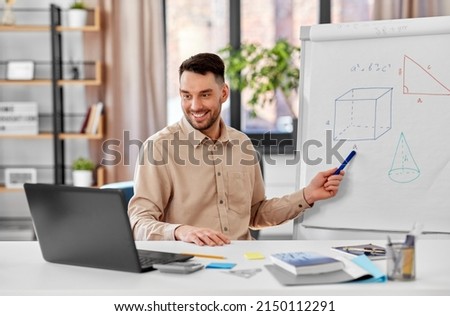 distant education, school and remote job concept - happy smiling male math teacher with laptop compute showing geometric shapes on flip chart having online class or video call at home office