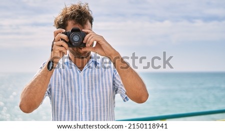 Young hispanic man smiling happy using camera at the beach.