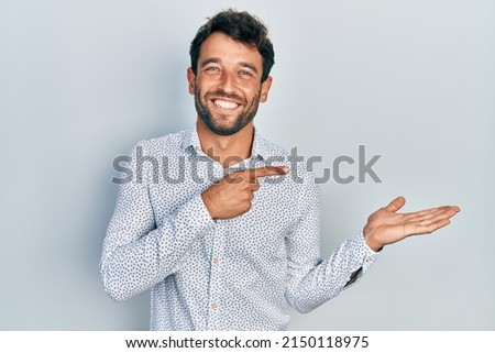 Handsome man with beard wearing casual elegant shirt amazed and smiling to the camera while presenting with hand and pointing with finger. 