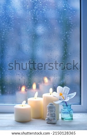 Buddha statue, candles and flower in cup on windowsill, abstract blurred rainy glass background. spring season. esoteric spiritual practice, Relax time, harmony, meditation, life balance concept.