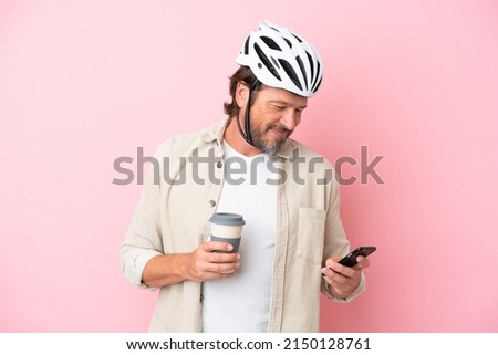 Senior dutch man with bike helmet isolated on pink background holding coffee to take away and a mobile