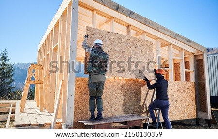 Carpenters mounting wooden OSB board on the wall of future cottage. Men workers building wooden frame house on pile foundation. Carpentry and construction concept. Royalty-Free Stock Photo #2150130359
