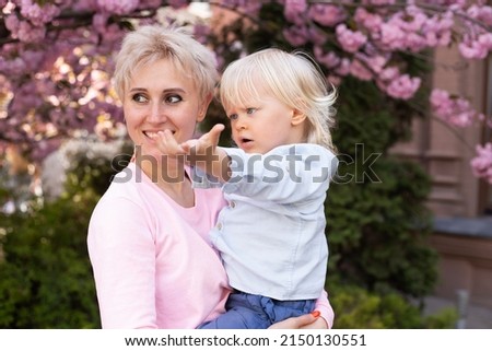 Young mother mom holding her little baby son boy child under blossoming Sakura Cherry trees with falling pink petals and beautiful flowers