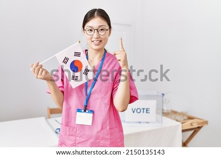 Young asian nurse woman at political campaign election holding south korea flag smiling with an idea or question pointing finger with happy face, number one 