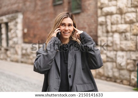 Young caucasian attractive smiling blonde woman in a grey jacket talking on a mobile phone flirting walking outside. Holding smartphone close to ear. Standing alone outside. Gloomy girl.  Royalty-Free Stock Photo #2150141633