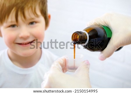 Happy little boy, smiling, wants to take the cure for the disease. Hands pouring cough medicine into a special measuring cap. Horizontal photo. Royalty-Free Stock Photo #2150154995