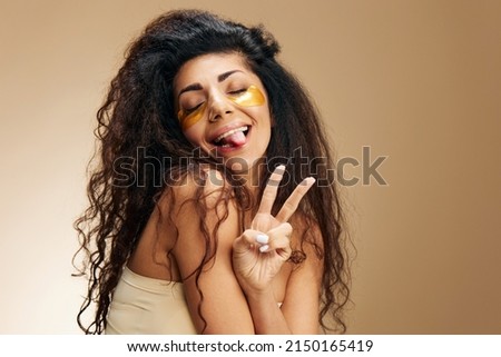 Cheerful Latin lady in gold hydrogel patches under closed eyes show v-sigh gesture stick tongue posing isolated over pastel beige background. Cosmetic Hair ad Natural beauty concept Studio Copy space