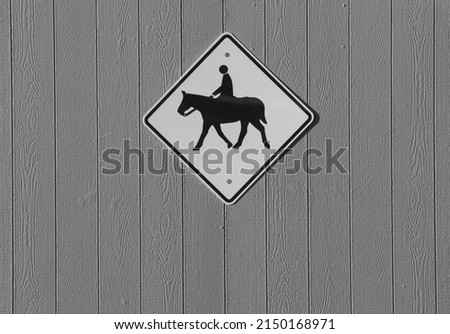 black and white of diamond shaped horse and rider warning sign on outside exterior barn wall signifying warning or beware or slow down due to horse back rider crossing horizontal format room for type
